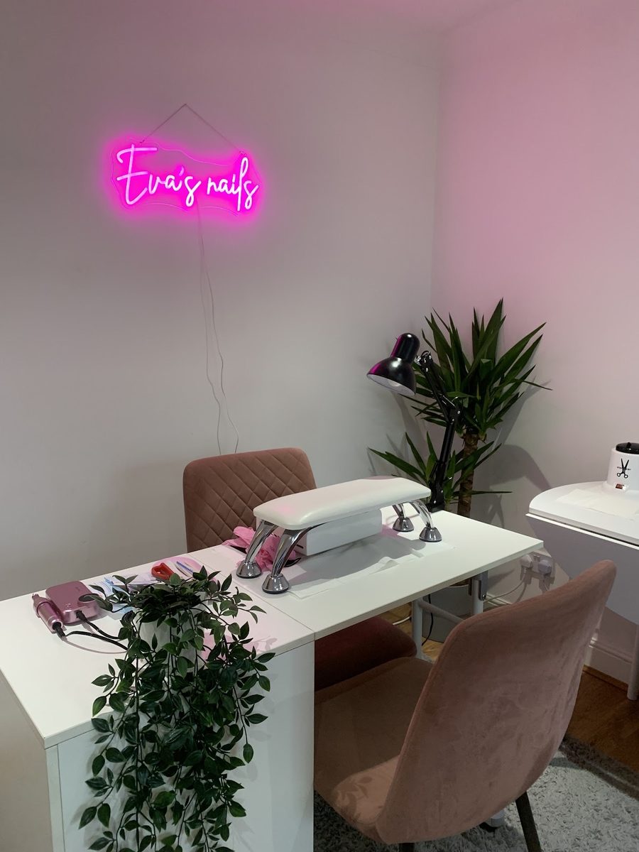 The salon interior: a white manicure table with dusty-pink chairs, a trailing plant and a pink neon sign on a pale pink wall